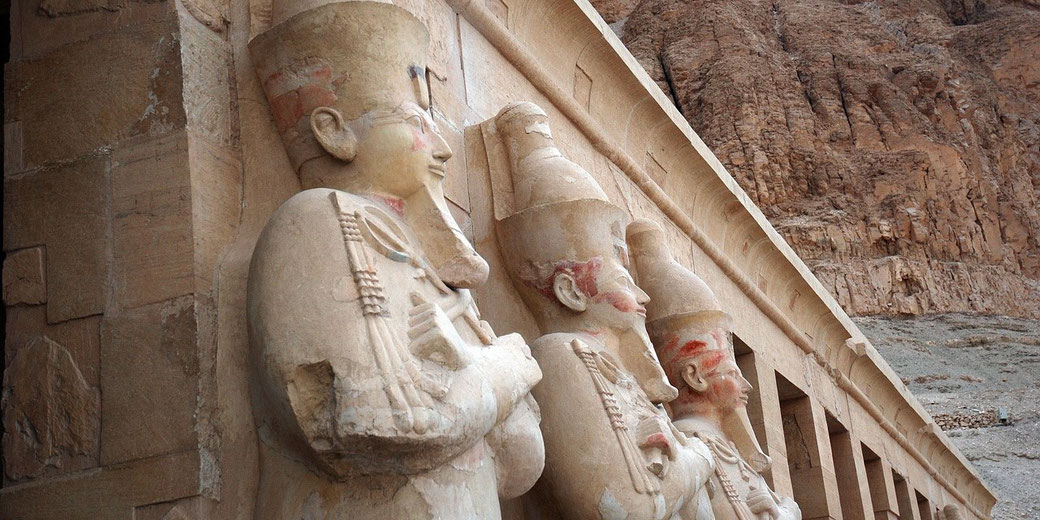 Row of ancient Egyptian statues with crossed arms and tall crowns, carved into a temple wall near a rocky cliff.