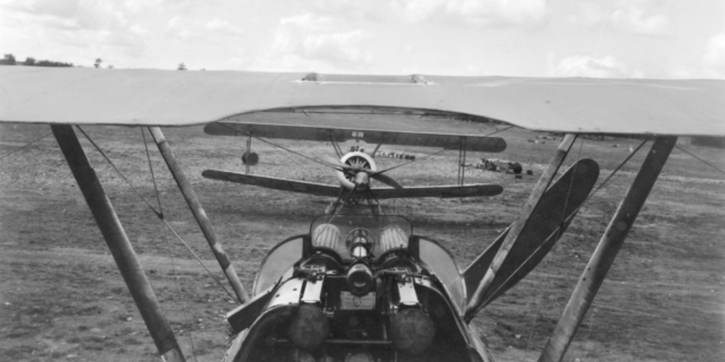 View from a biplane's gunner seat showing twin machine guns and a second biplane directly ahead on a dirt airfield.