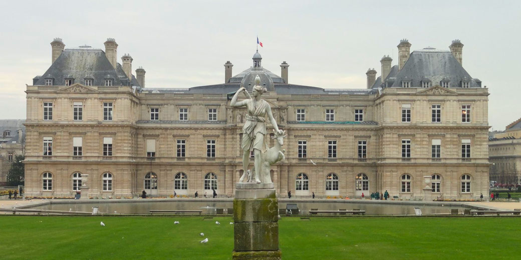 A classical statue stands before the grand Luxembourg Palace in Paris, with the French flag atop and green lawn in the foreground.