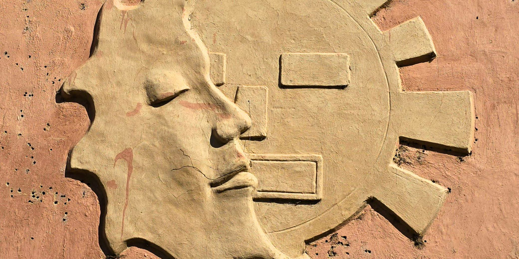 A bas-relief of a stylized human face in profile, integrated with a geometric sun design.