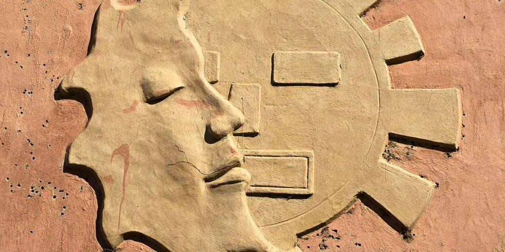 A bas-relief of a stylized human face in profile, integrated with a geometric sun design.