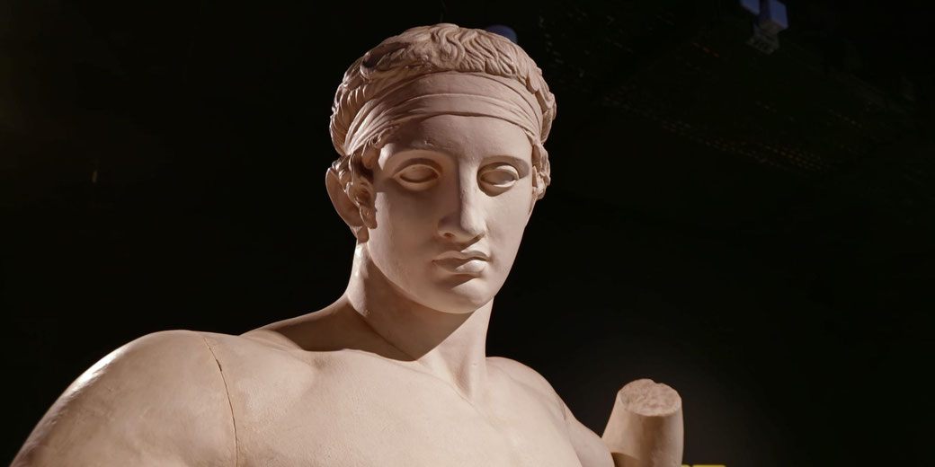 A classical Greek statue of a male athlete, possibly a victor, with a headband. The finely sculpted details and lifelike expression reflect ancient Greek artistry and athletic ideals.