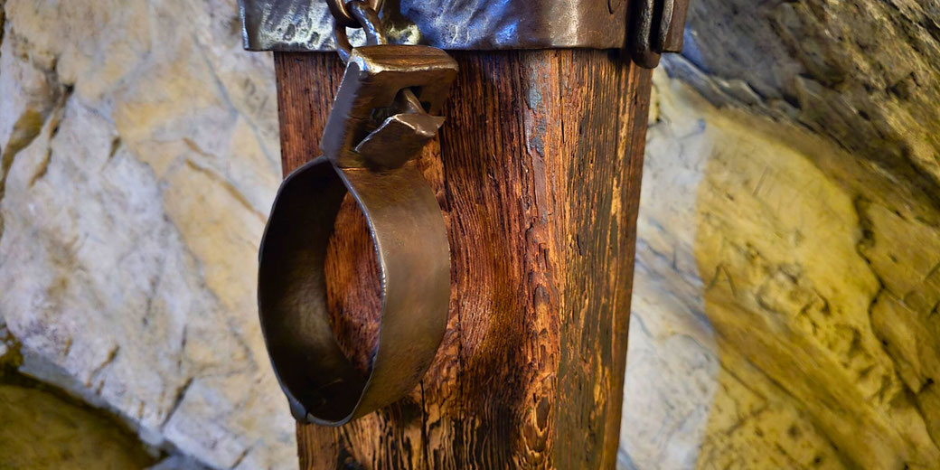 A medieval iron shackle hangs from a weathered wooden post, set against a rough stone wall. The rusted metal and aged wood evoke a sense of historical imprisonment or punishment.
