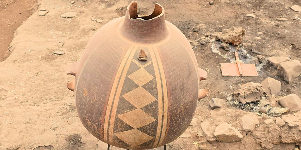 A large, round, ancient clay pot with geometric patterns, a broken rim, and side protrusions sits in an archaeological excavation site with scattered stones and debris in the background.