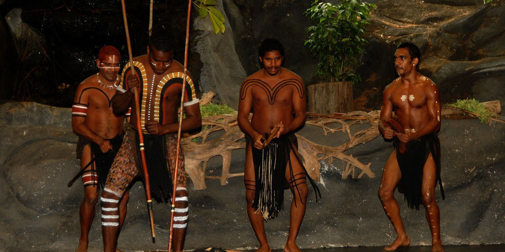 Four Indigenous Australian men perform a traditional dance, wearing body paint and cultural attire. They hold sticks and spears.