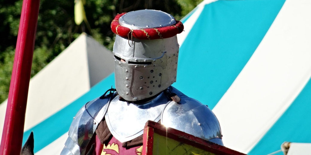 A medieval knight in silver armour stands near striped tents at an outdoor fair or reenactment event in daylight.