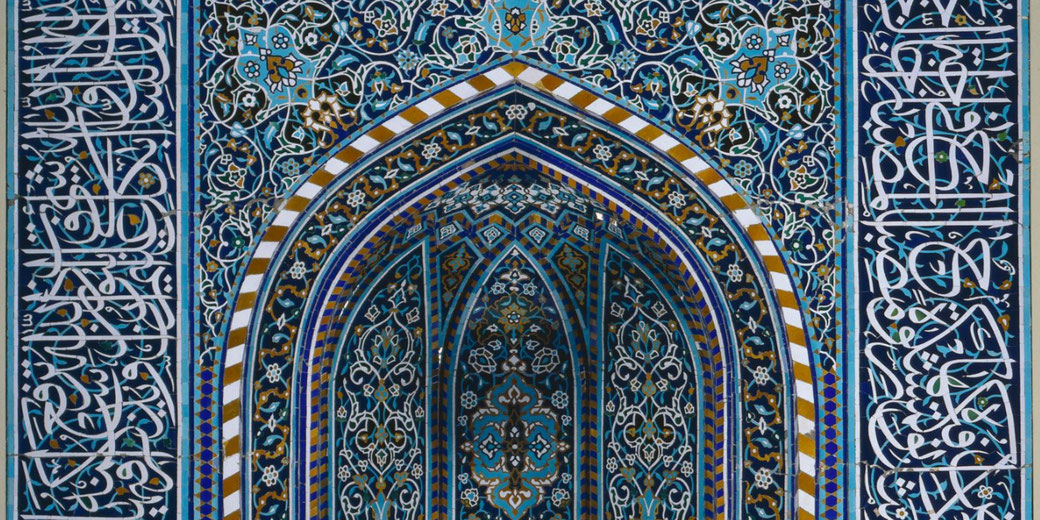 Close-up of Islamic geometric and floral tilework with Arabic calligraphy, featuring vivid blue, gold, and white patterns in a pointed arch design.