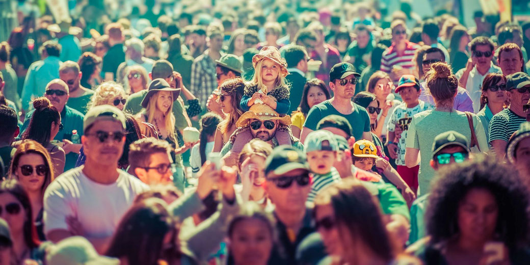 A vibrant crowd of diverse people fills the scene, with a young girl sitting on a man's shoulders, both wearing straw hats.