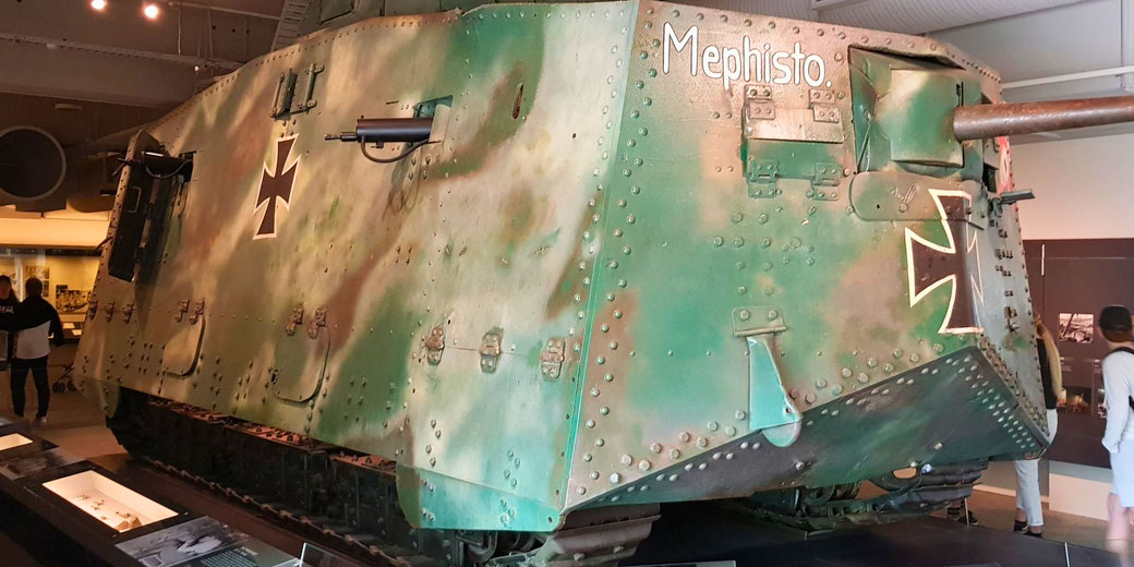 A large, green WWI German A7V tank named "Mephisto" on display in a museum, featuring riveted armor, guns on the sides, and black Iron Cross insignias.