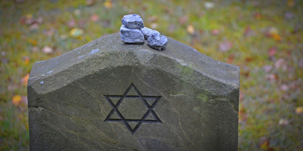 A Jewish headstone marked with the Star of David has small memorial stones placed on top, set against a grassy background with scattered leaves.