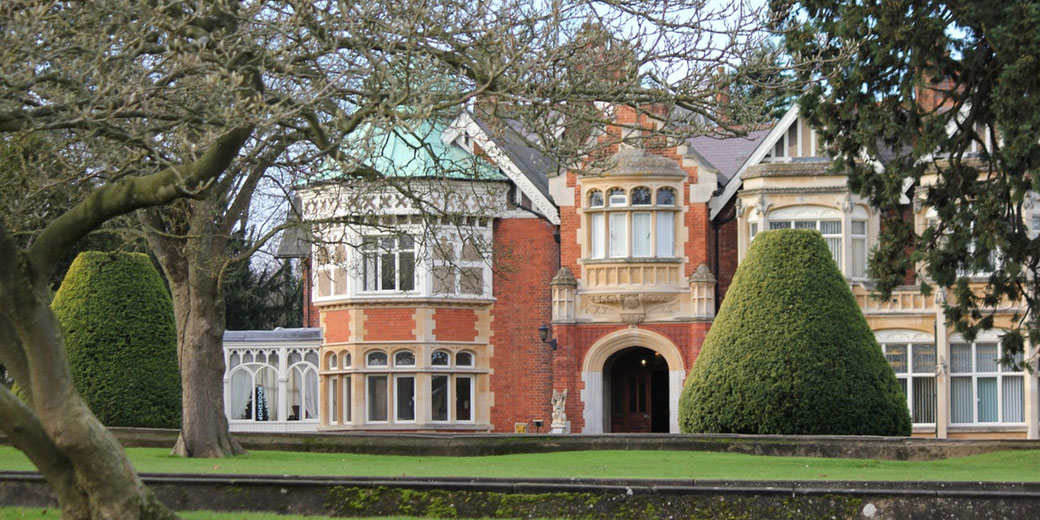 A large brick mansion with a green copper roof sits behind trimmed trees and hedges on a well-kept lawn.