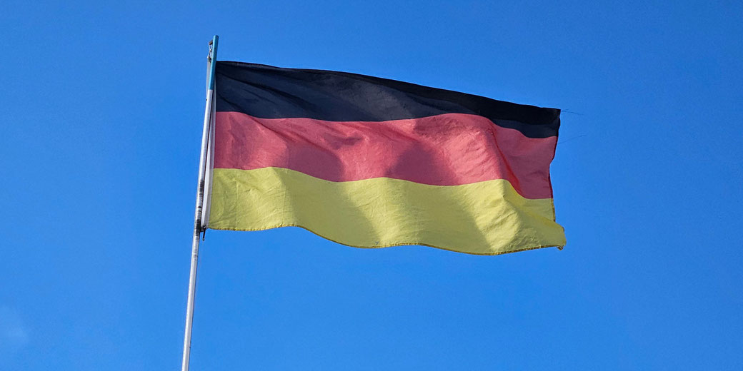 A German flag with black, red, and gold stripes waves against a clear blue sky.