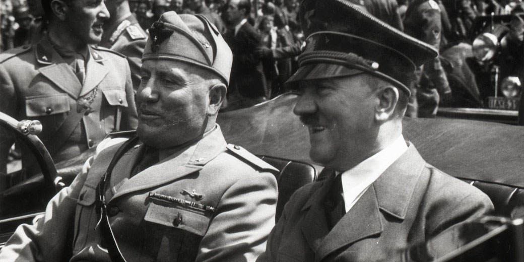 Adolf Hitler and Benito Mussolini sit together in an open car, smiling, surrounded by uniformed men and a crowd in the background.