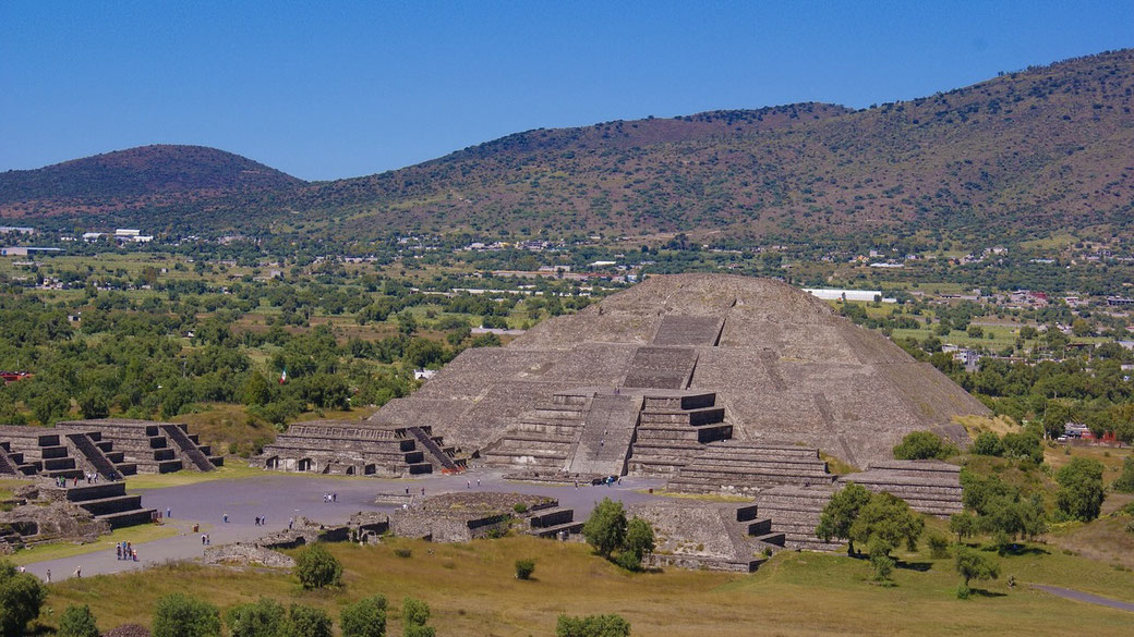 Pyramid of the Sun at Teotihuacan, Mexico, an ancient Mesoamerican structure. The massive stone pyramid is surrounded by smaller ruins, with green hills and a blue sky in the background.