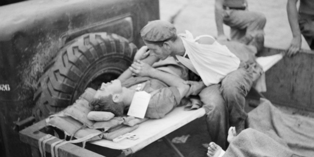 Black and white photo of a wounded soldier on a stretcher being comforted by another injured man with a bandaged arm, beside a military vehicle.