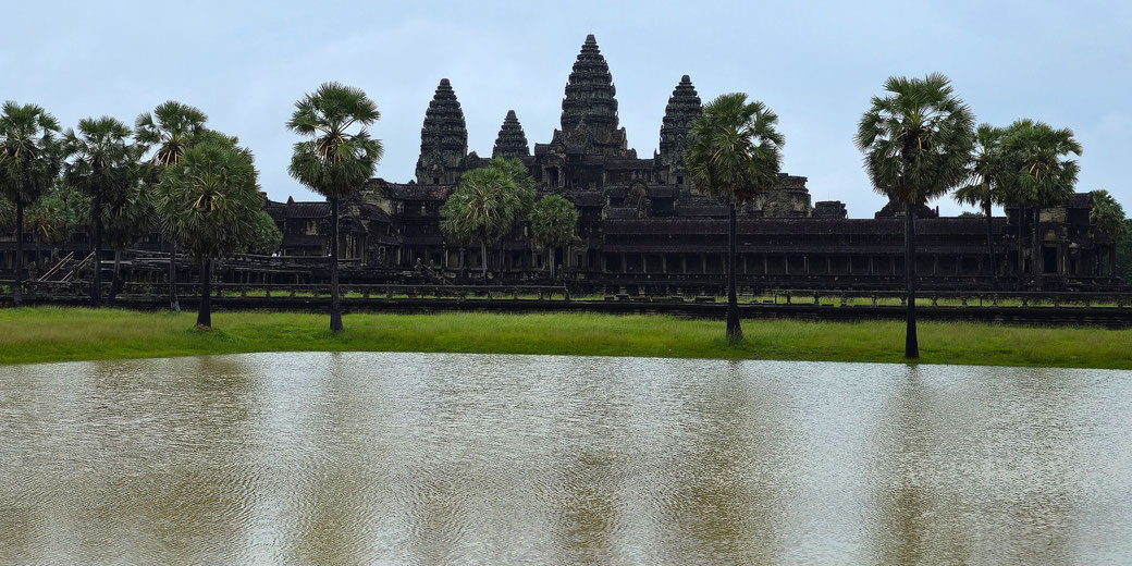 Angkor Wat temple with its towers rising behind palm trees, seen across a pond with calm water and green grass.