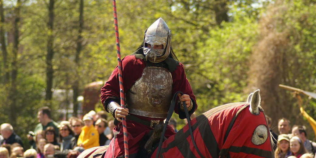 Armored knight on horseback holds a lance during a medieval reenactment, surrounded by spectators in a wooded outdoor setting.
