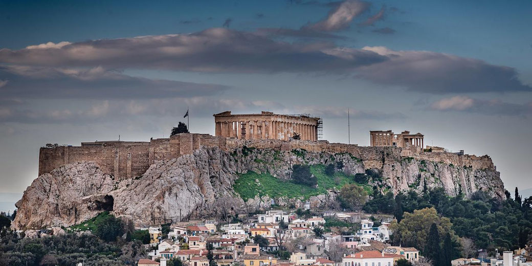 The Acropolis of Athens rises above the city with the Parthenon and other ruins atop the rocky hill under a cloudy sky.
