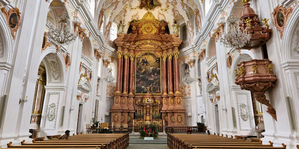 The ornate interior of a Baroque-style church, featuring intricate white and gold decorations, chandeliers, a grand altar, and painted ceilings, with rows of wooden pews.