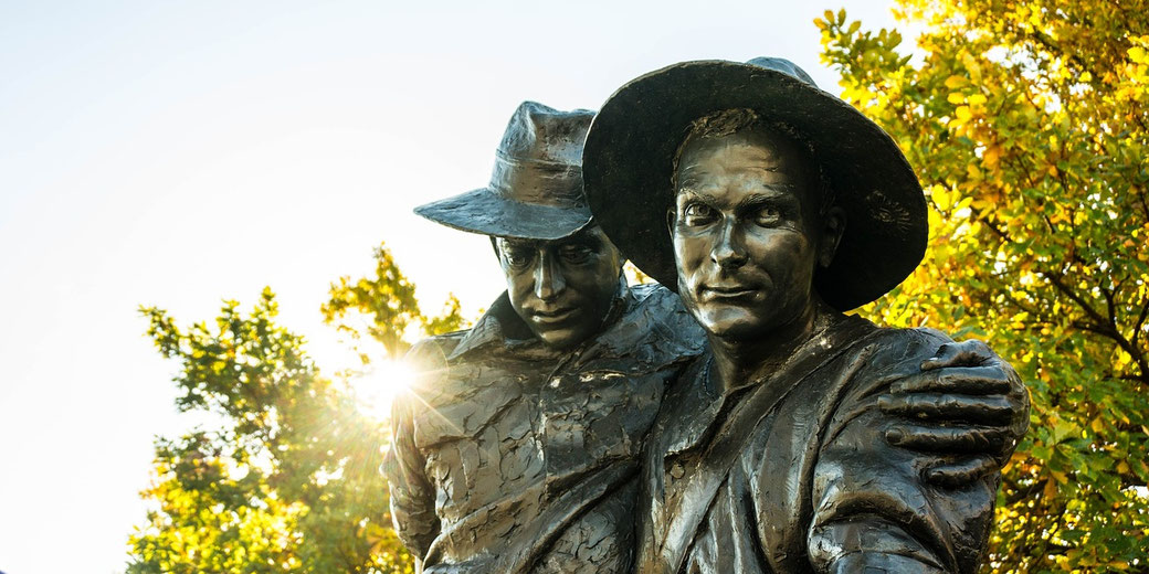 A bronze statue shows one soldier supporting another, set against sunlit trees and a bright sky.