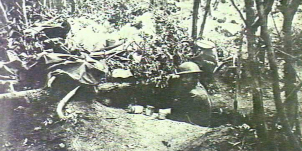 Black and white photo of soldiers in a camouflaged jungle trench, partially hidden by foliage and gear, with helmets and supplies visible.