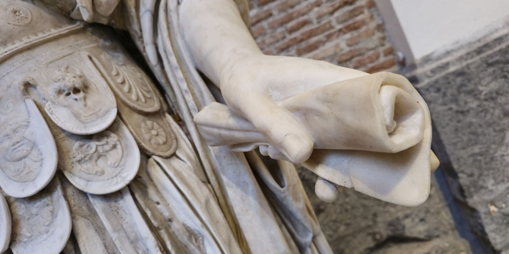 Close-up of a marble statue's hand holding a scroll, with detailed armor featuring faces and decorative patterns visible in the background.