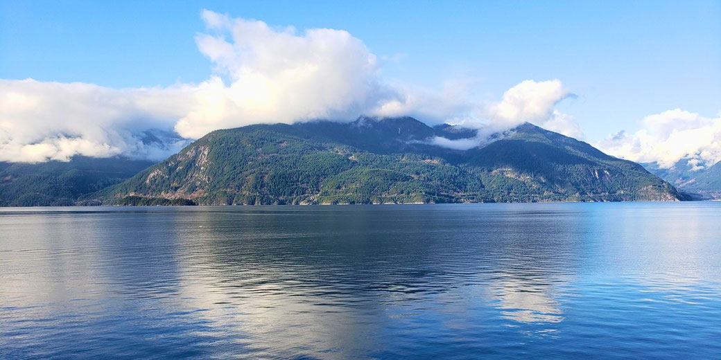 A serene lake reflects lush green mountains partially covered by soft, white clouds. The blue sky and calm waters create a peaceful, picturesque landscape.