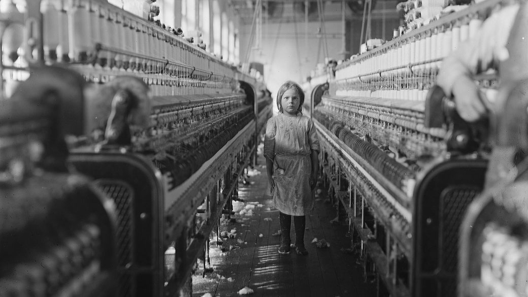 A young girl in a worn dress stands barefoot between long rows of spinning machines in a textile mill.