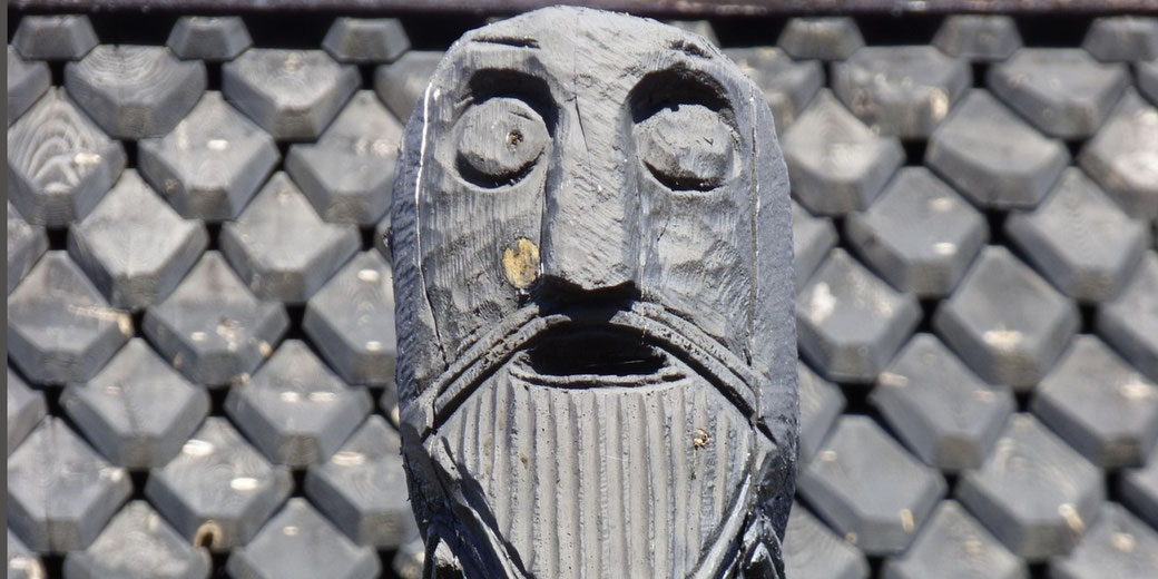 A carved wooden face with deep-set eyes and a long beard stands against a wall of similarly shaped, hexagonal wooden blocks.