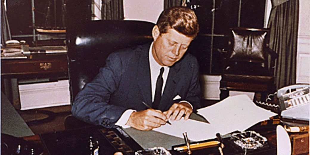 John F. Kennedy sits at a desk, writing on a document with a pen, in a formal office setting.