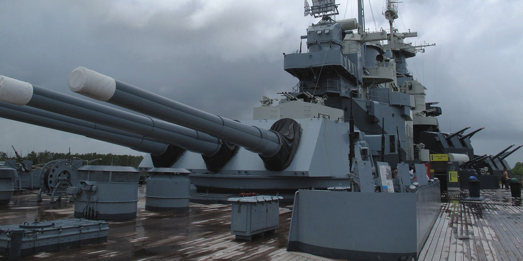 A large naval battleship with triple gun turrets and radar towers is docked under a cloudy sky, viewed from its wooden deck.