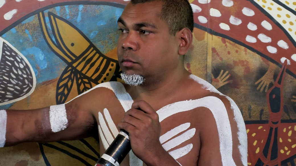 A man with traditional white body paint and a painted beard speaks into a microphone, standing against a vibrant Indigenous Australian artwork featuring animals and handprints.