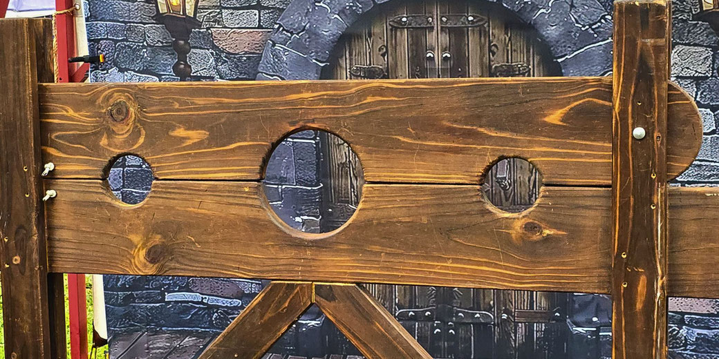 A wooden medieval-style stockade with three circular holes for securing a person's head and hands.