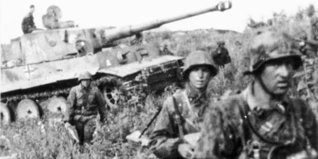 Soldiers in camouflage gear move through tall grass near a heavily armed tank, with one crew member visible on top of the vehicle.