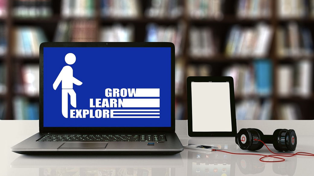 A laptop displaying "GROW LEARN EXPLORE," alongside a tablet, headphones, and a USB drive on a desk, with bookshelves in the background, symbolizing digital learning and education.