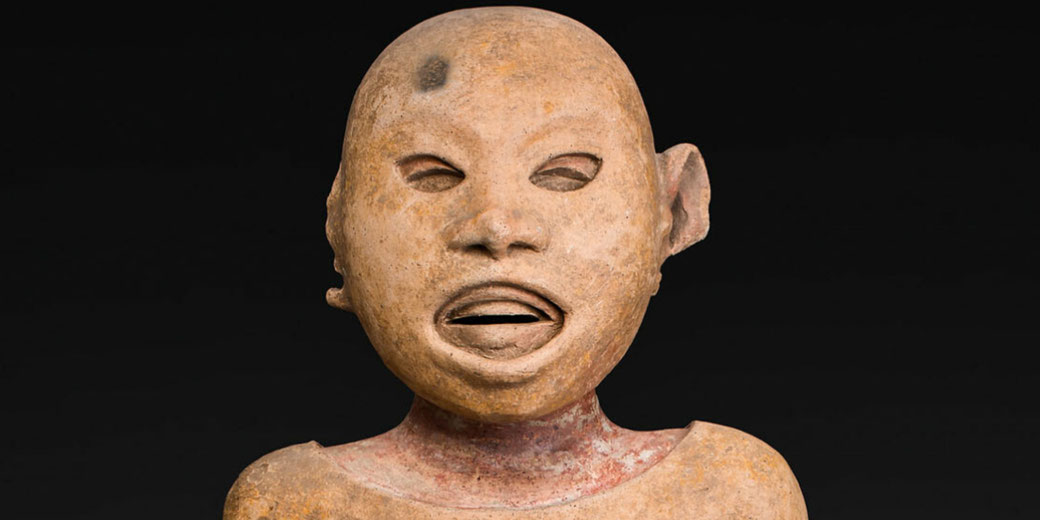 A terracotta sculpture of a human figure with a rounded head, hollowed eyes, an open mouth, and a slightly distorted ear. The weathered surface has earthy tones and traces of red pigment on the neck.