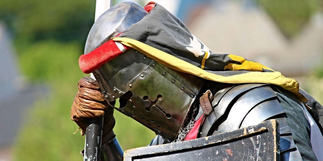 A medieval knight in full armor leans on his sword, wearing a detailed helmet with a cross-shaped visor and a colorful fabric draped over it.