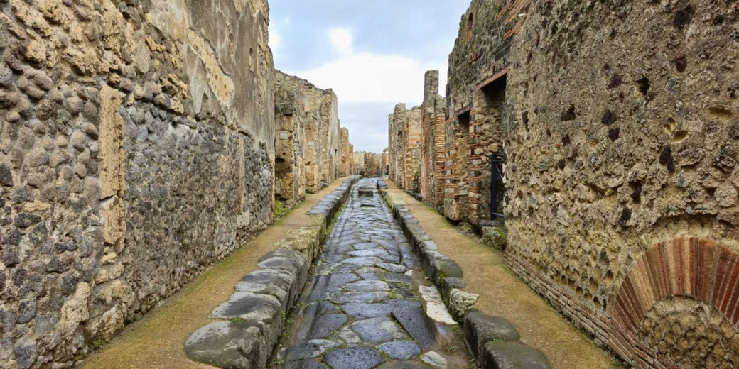 Ancient stone street flanked by weathered stone and brick walls, showing the ruins of a once-thriving Roman city under an overcast sky.