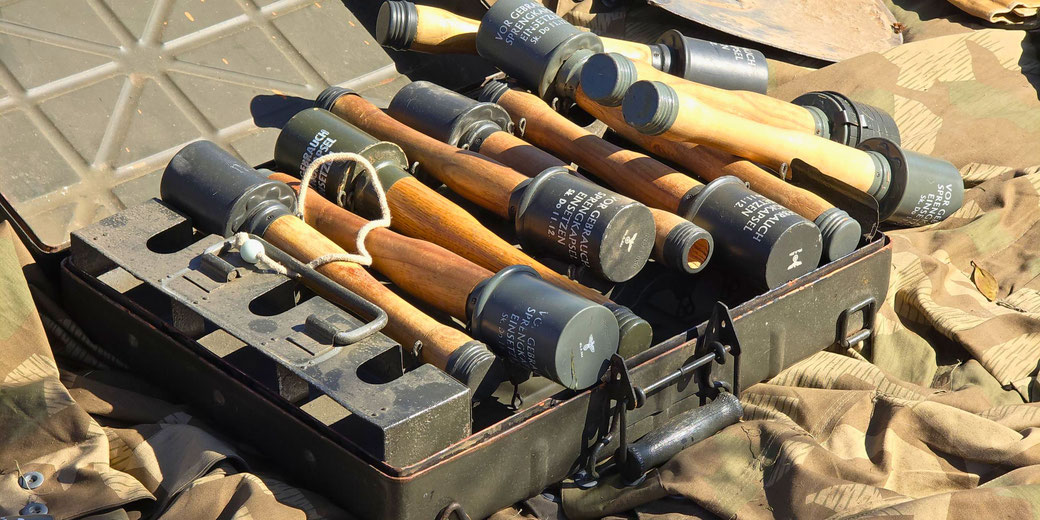 A metal crate filled with German stick grenades rests on camouflage fabric, with wooden handles and labeled metal heads visible.