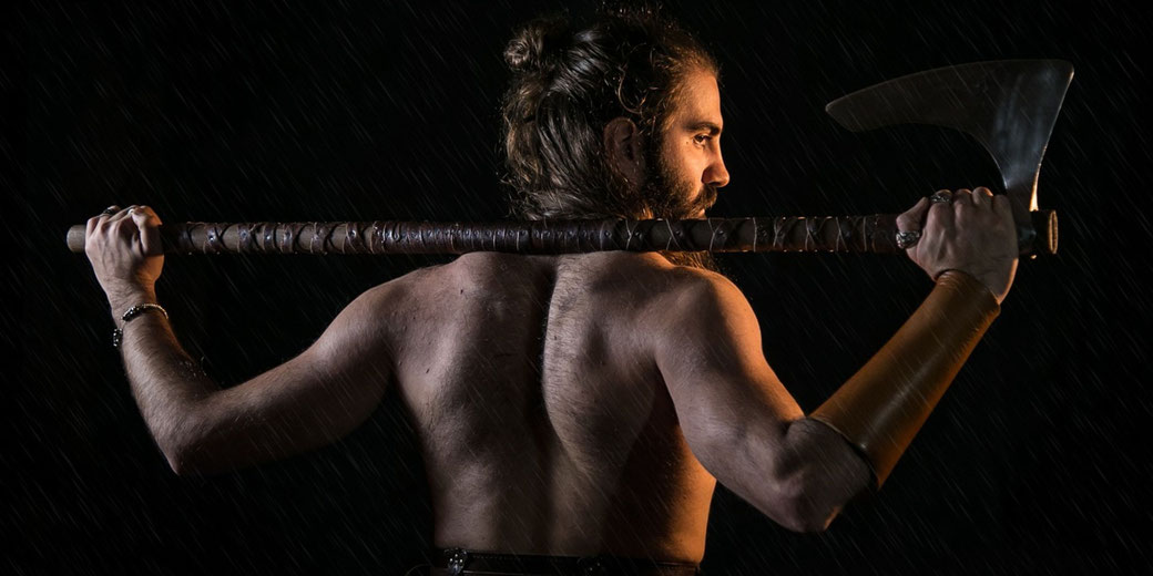 A muscular man with long hair holds a large axe across his shoulders, standing shirtless against a dark background.