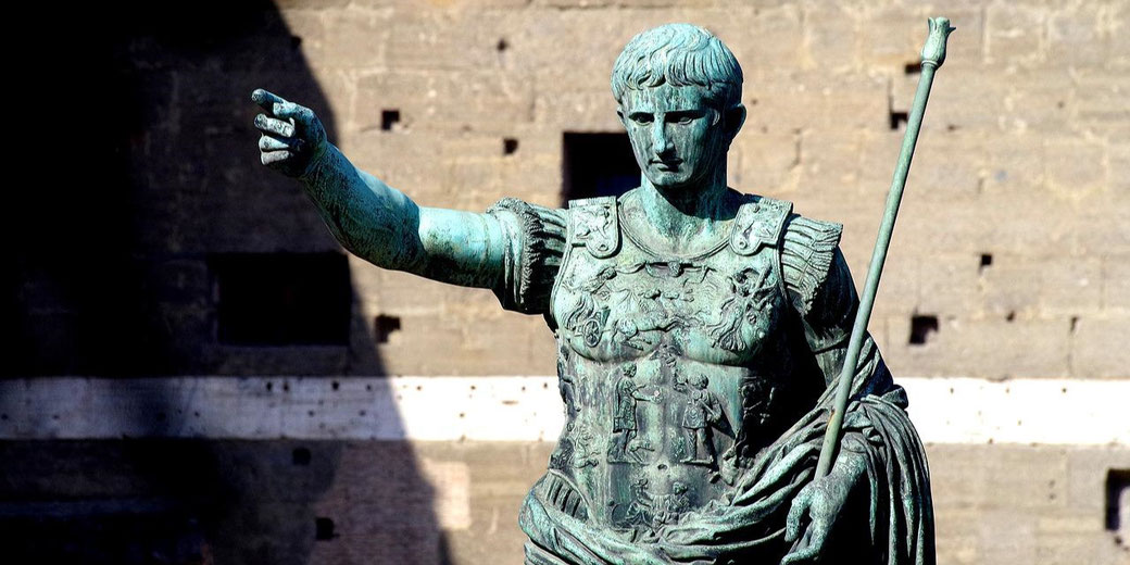 Bronze statue of a Roman emperor in detailed armor, pointing forward with a staff in the other hand.