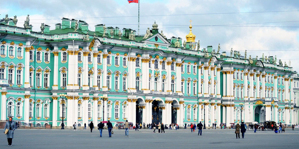 Grand pastel-green and white palace with gold accents, columns, and statues, facing a large square filled with people.