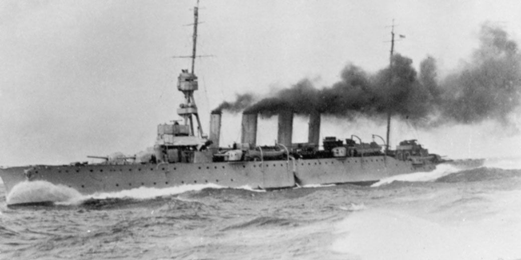 A warship with four smokestacks moves through rough seas, emitting thick black smoke as it sails at speed.