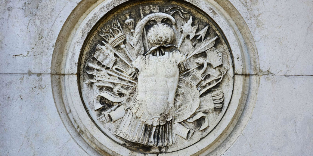 An intricate stone relief depicting a headless suit of armor surrounded by weapons, banners, and military symbols. The weathered carving suggests a historical or commemorative theme, rich in detail and texture.