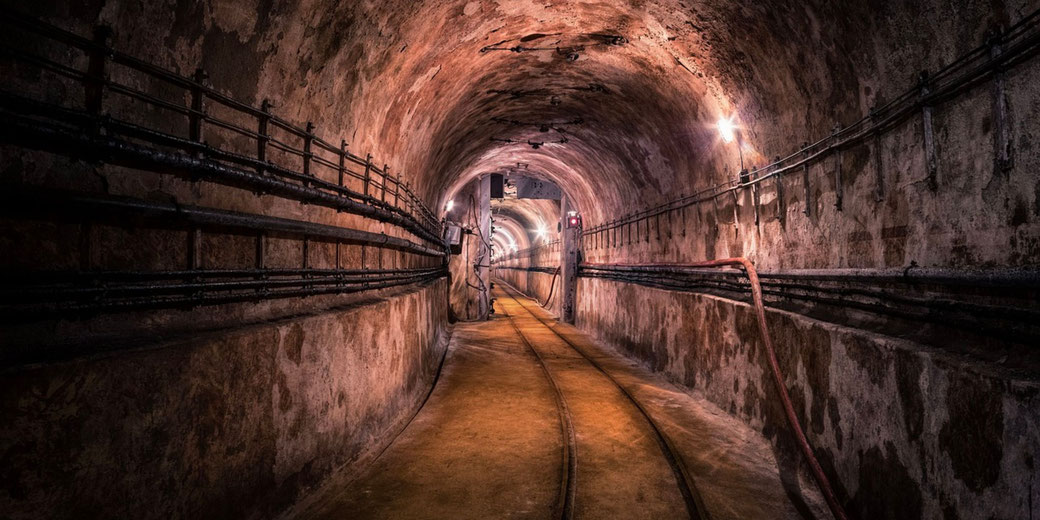 A dimly lit, aged underground tunnel with rusty pipes and narrow railway tracks curving into the distance.