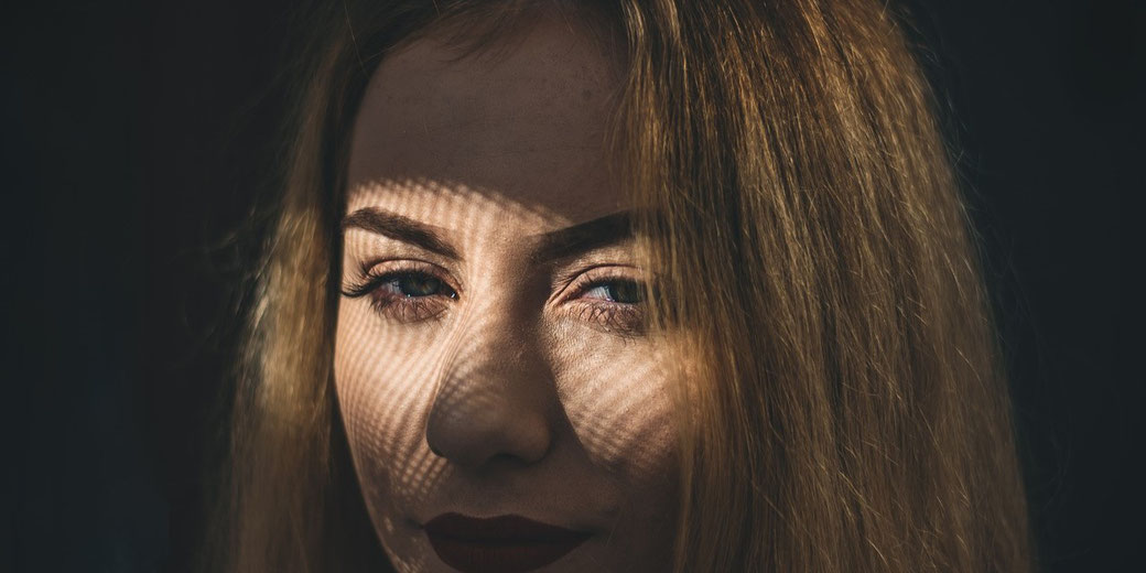 Close-up of a woman's face with dramatic shadow patterns from blinds across her eyes, highlighting her intense gaze and red lipstick.