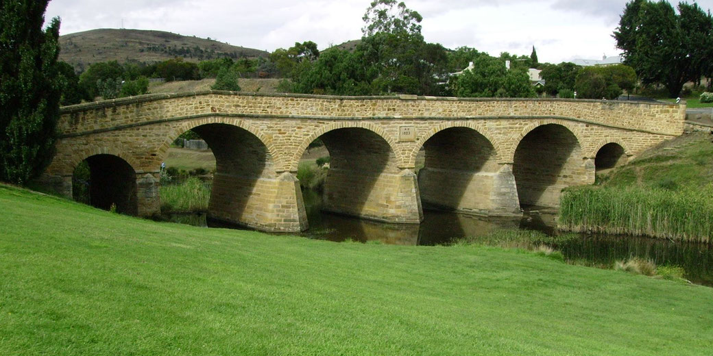A stone arch bridge crosses a narrow river, surrounded by grass and trees, with hills in the background.