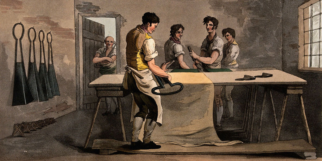 A group of workers in a dimly lit workshop are shearing cloth. Large shears hang on the wall, and a man in the foreground holds a pair while cutting fabric.