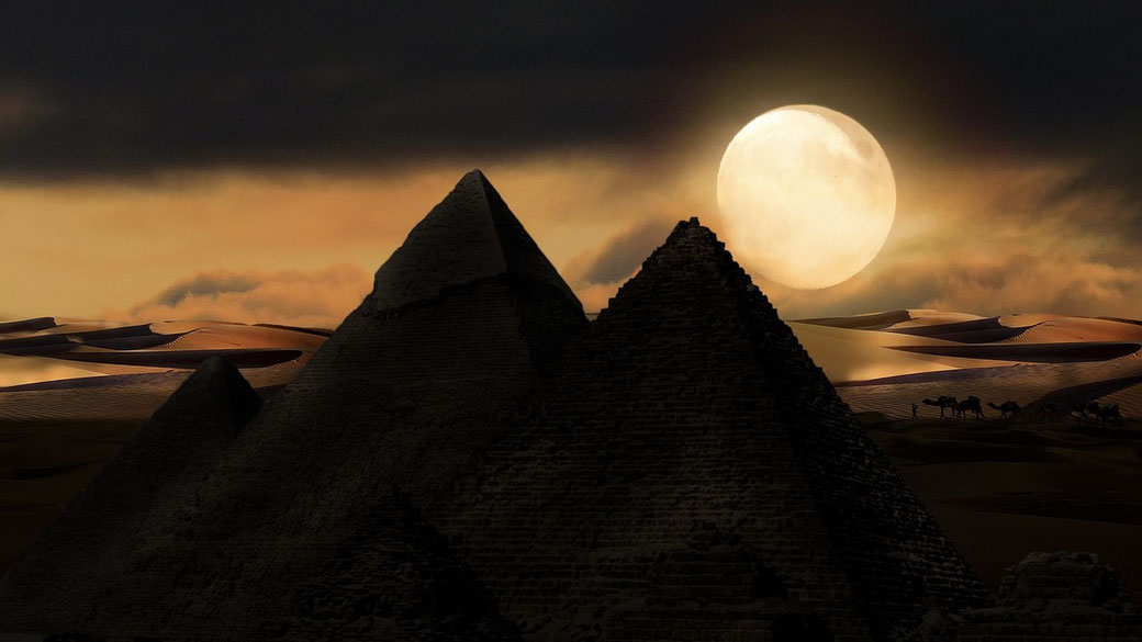 The Pyramids of Giza at night, silhouetted against a dramatic sky with a glowing full moon. Sand dunes and a caravan of camels add to the mystical desert atmosphere.