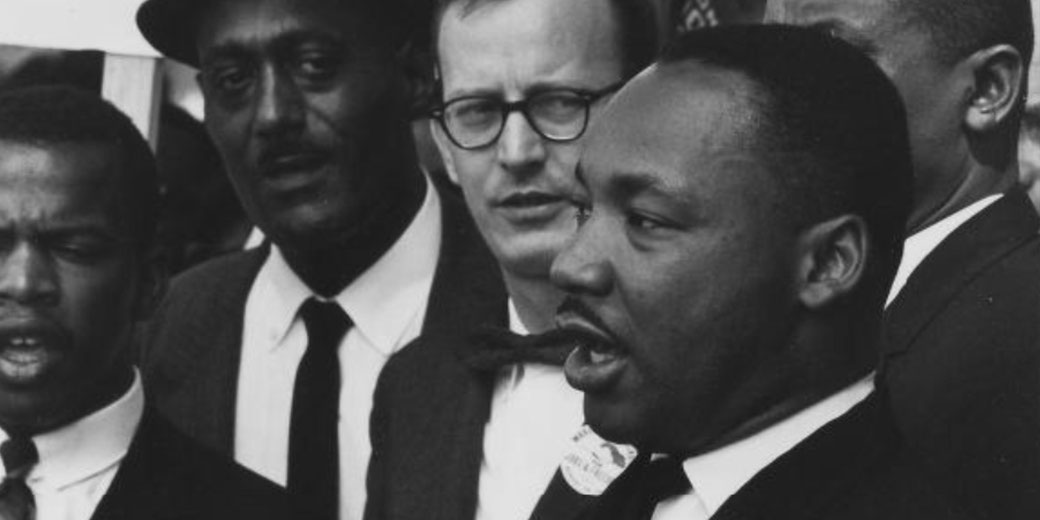 Black-and-white photo of Martin Luther King Jr. speaking with a group of civil rights leaders, all dressed in suits, appearing focused and determined.